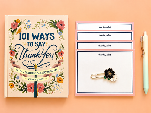 A flat-lay of a stationery set on a peach background, featuring a hardcover book titled '101 Ways to Say Thank You' by Kelly Browne, matching note cards, a pen, and a flower-themed paperclip. Curated by Amazing Robin Gifts.
