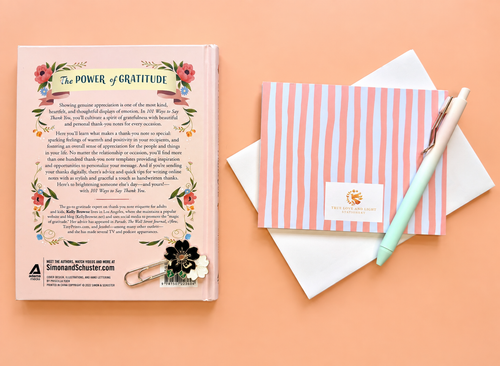 A styled flat-lay of a book titled 'The Power of Gratitude' showing its back cover, next to a pink and white striped envelope, a white card, and a mint green pen on a peach background. This the back image of the 101 Ways to Say Thank You Book & Cards Gift Set, curated by Amazing Robin Gifts.