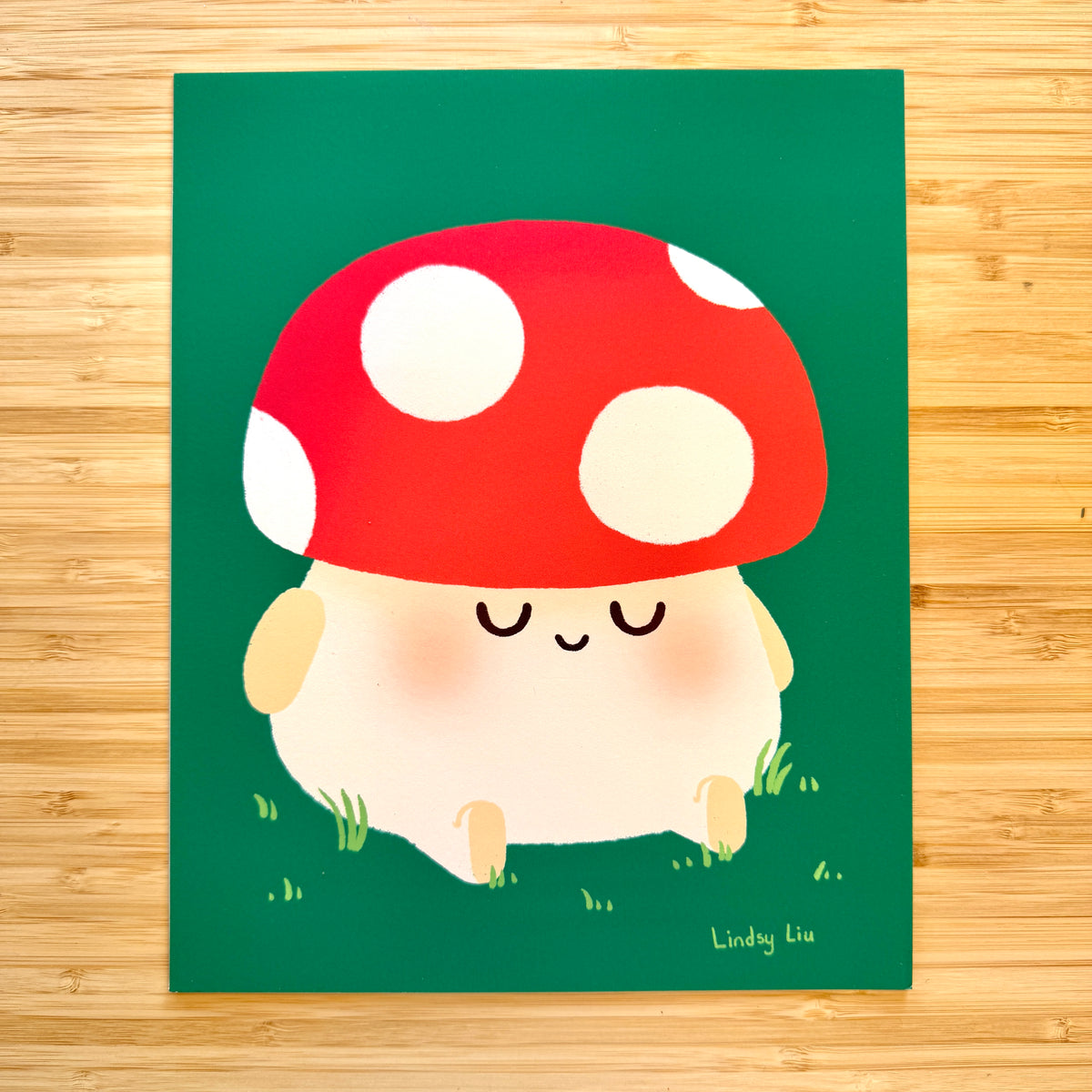 A square art print featuring a cute cartoon mushroom character with a red cap, white spots, and a cream body, placed on a green background and photographed flat on a light wood surface. The artist signature 'Lindsy Liu' is visible in the bottom right corner of the artwork.