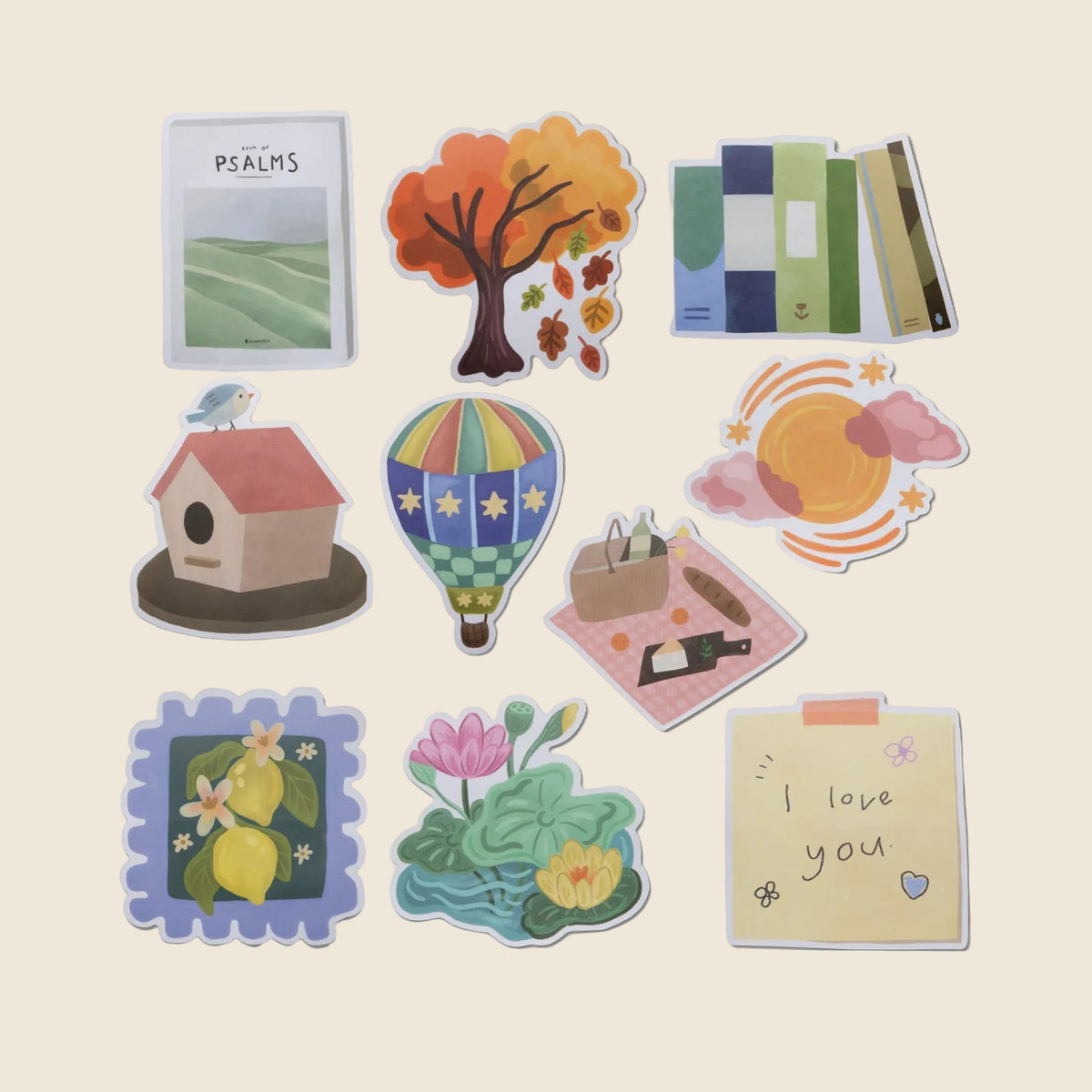A set of nine die-cut sticker illustrations arranged on a light beige background. The stickers feature whimsical designs including a tree with autumn leaves, a hot air balloon, a birdhouse, lemons, a lotus pond, a picnic, a notebook page reading 'I love you', and books. Designed by Alabaster Co. 