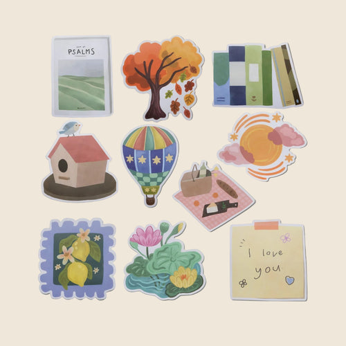 A set of nine die-cut sticker illustrations arranged on a light beige background. The stickers feature whimsical designs including a tree with autumn leaves, a hot air balloon, a birdhouse, lemons, a lotus pond, a picnic, a notebook page reading 'I love you', and books. Designed by Alabaster Co. 