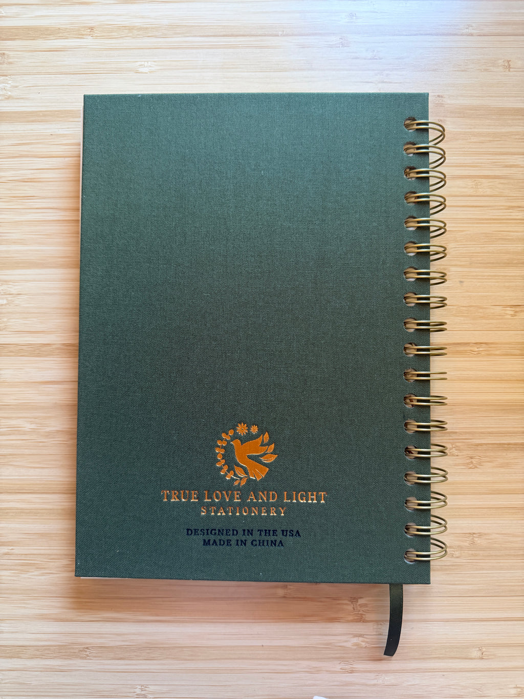 A dark green spiral-bound notebook with a gold wire binding and a black ribbon bookmark, placed on a light wood surface. The cover features a gold logo and the text 'TRUE LOVE AND LIGHT STATIONERY'.