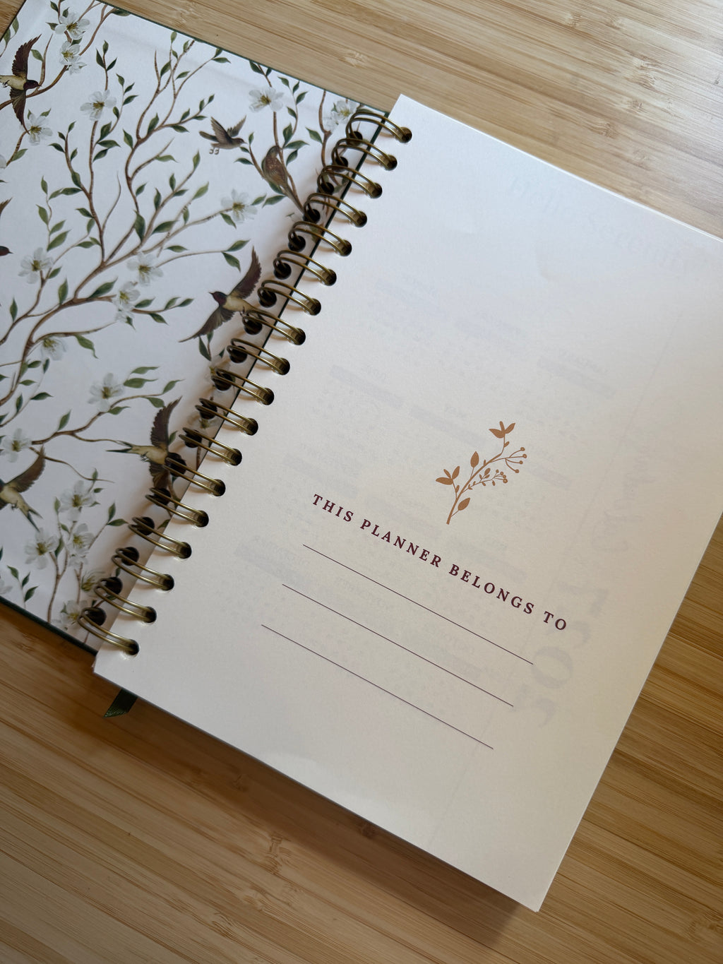An open spiral-bound planner lying on a light wood surface. The left page shows a white cover with a delicate botanical and bird illustration in muted green, brown, and gray tones.