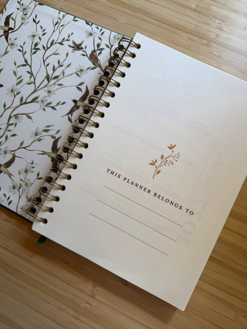An open spiral-bound planner lying on a light wood surface. The left page shows a white cover with a delicate botanical and bird illustration in muted green, brown, and gray tones.