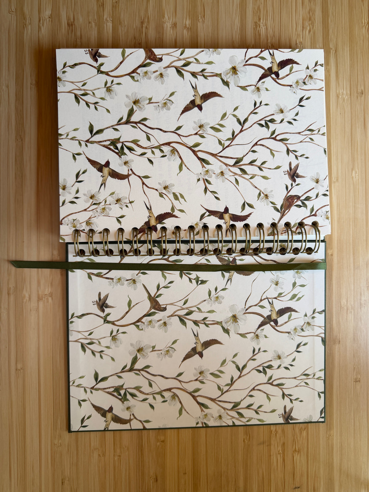 A spiral-bound notebook with a cream-colored cover decorated with a detailed pattern of hummingbirds, branches, and white blossoms, displayed flat on a light wood background.