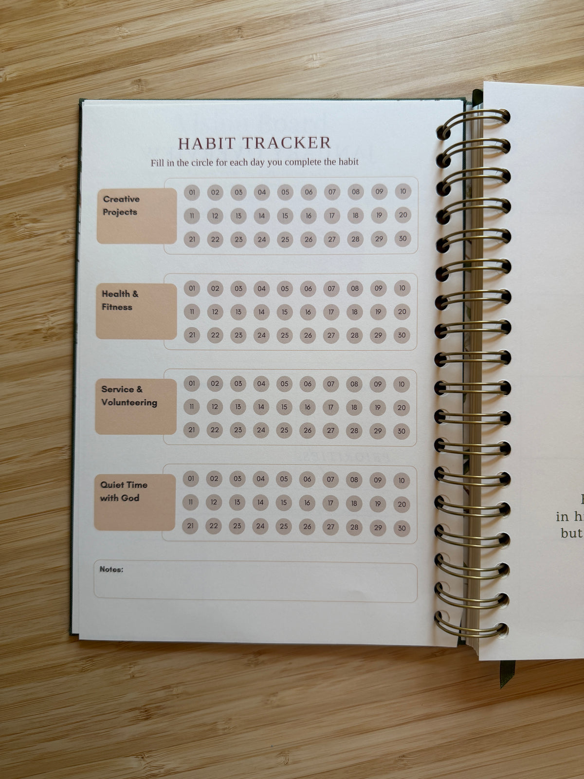 An open spiral-bound habit tracker notebook on a light wood surface. The page shows labeled boxes for Creative Projects, Health & Fitness, Service & Volunteering, and Quiet Time with God, each with rows of small circles for daily tracking.