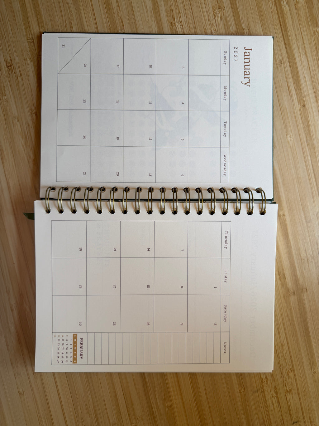 An open spiral-bound planner or calendar showing blank monthly calendar pages on a light wood surface.