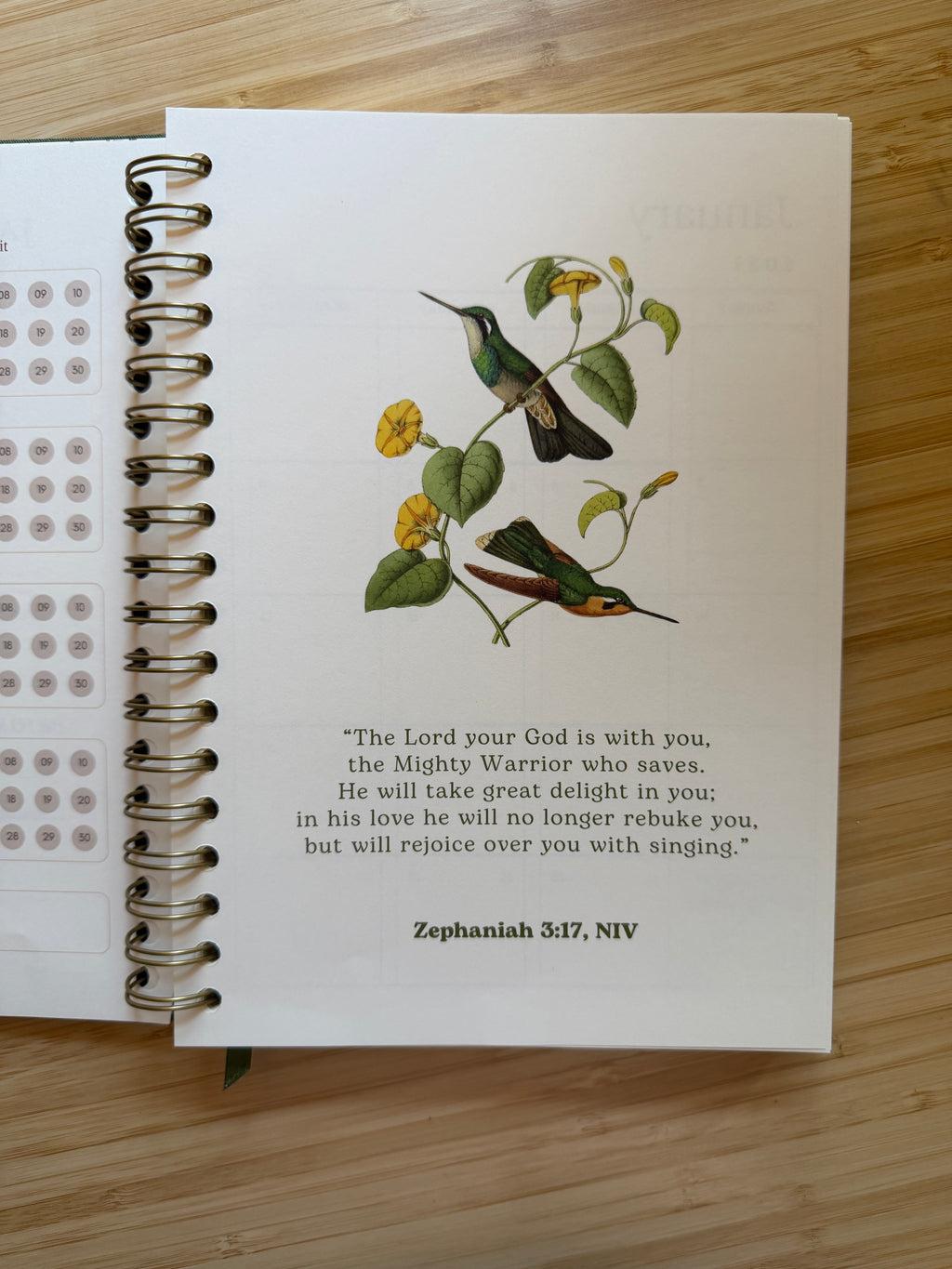 An open spiral-bound journal or planner lying on a light wood surface, showing a printed page with two illustrated birds, small yellow flowers, a Bible verse, and the heading 'Zephaniah 3:17, NIV.'.