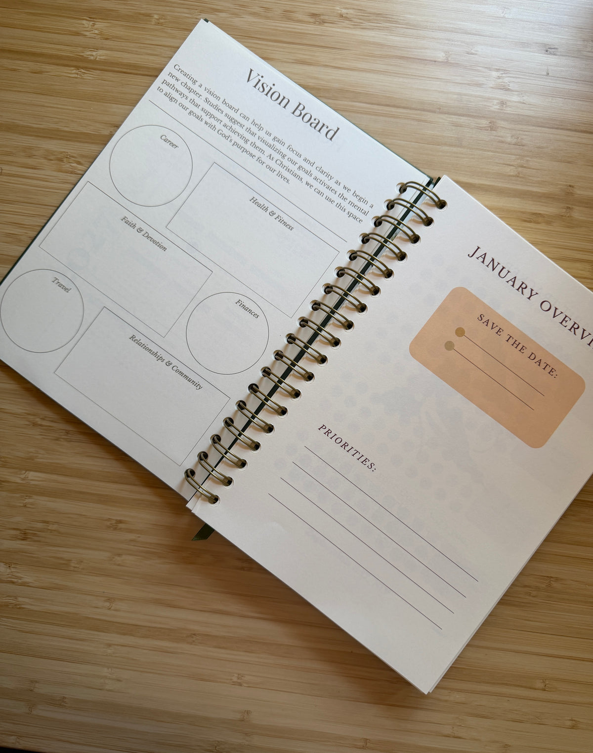 An open spiral-bound journal lying on a light wood surface, showing a Vision Board and January Overview page with lined boxes and a tan 'Save the Date' box.
