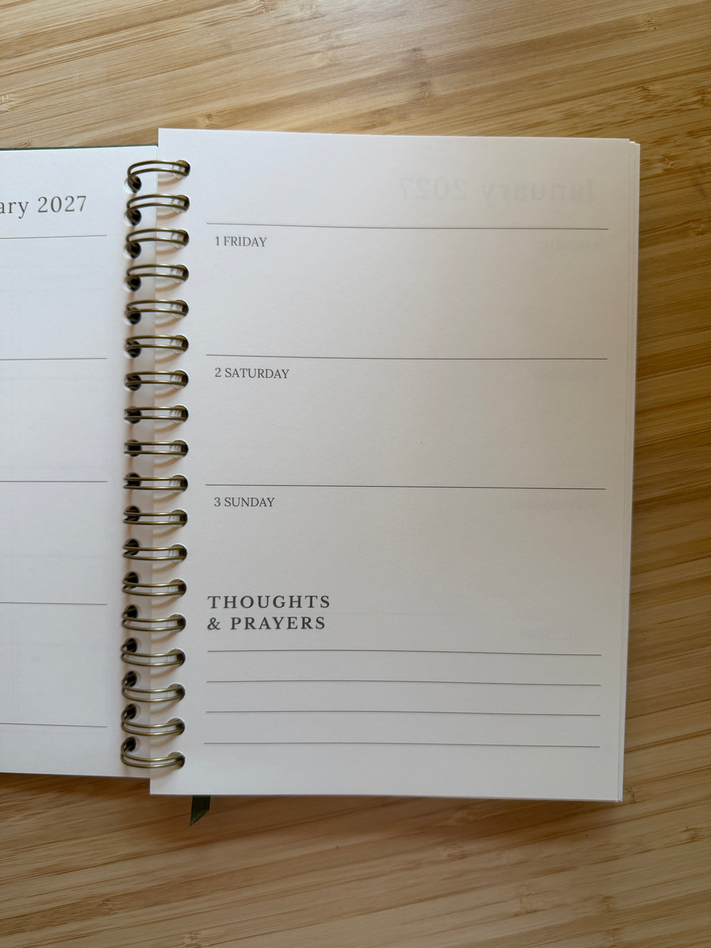 An open spiral-bound planner or journal lying on a light wood surface, showing a weekly layout with sections for Friday through Sunday and a 'THOUGHTS & PRAYERS' column.