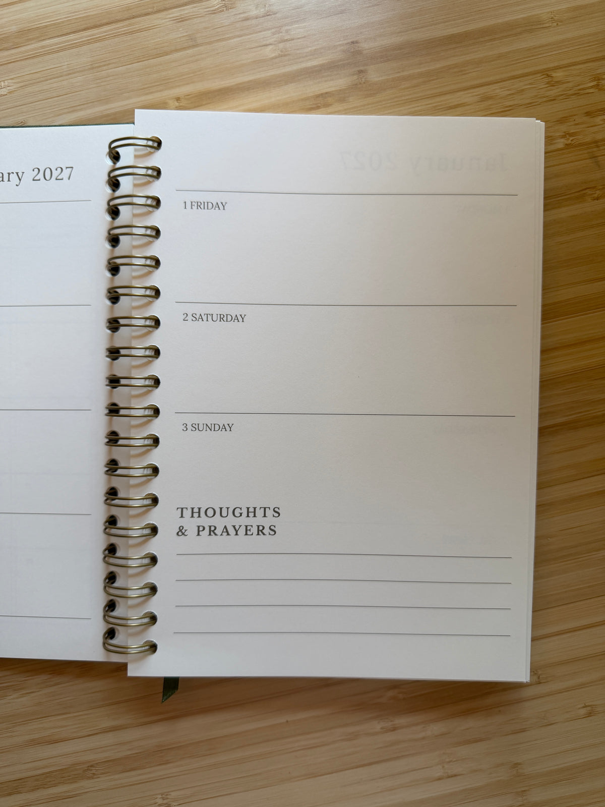 An open spiral-bound planner or journal lying on a light wood surface, showing a weekly layout with sections for Friday through Sunday and a 'THOUGHTS & PRAYERS' column.