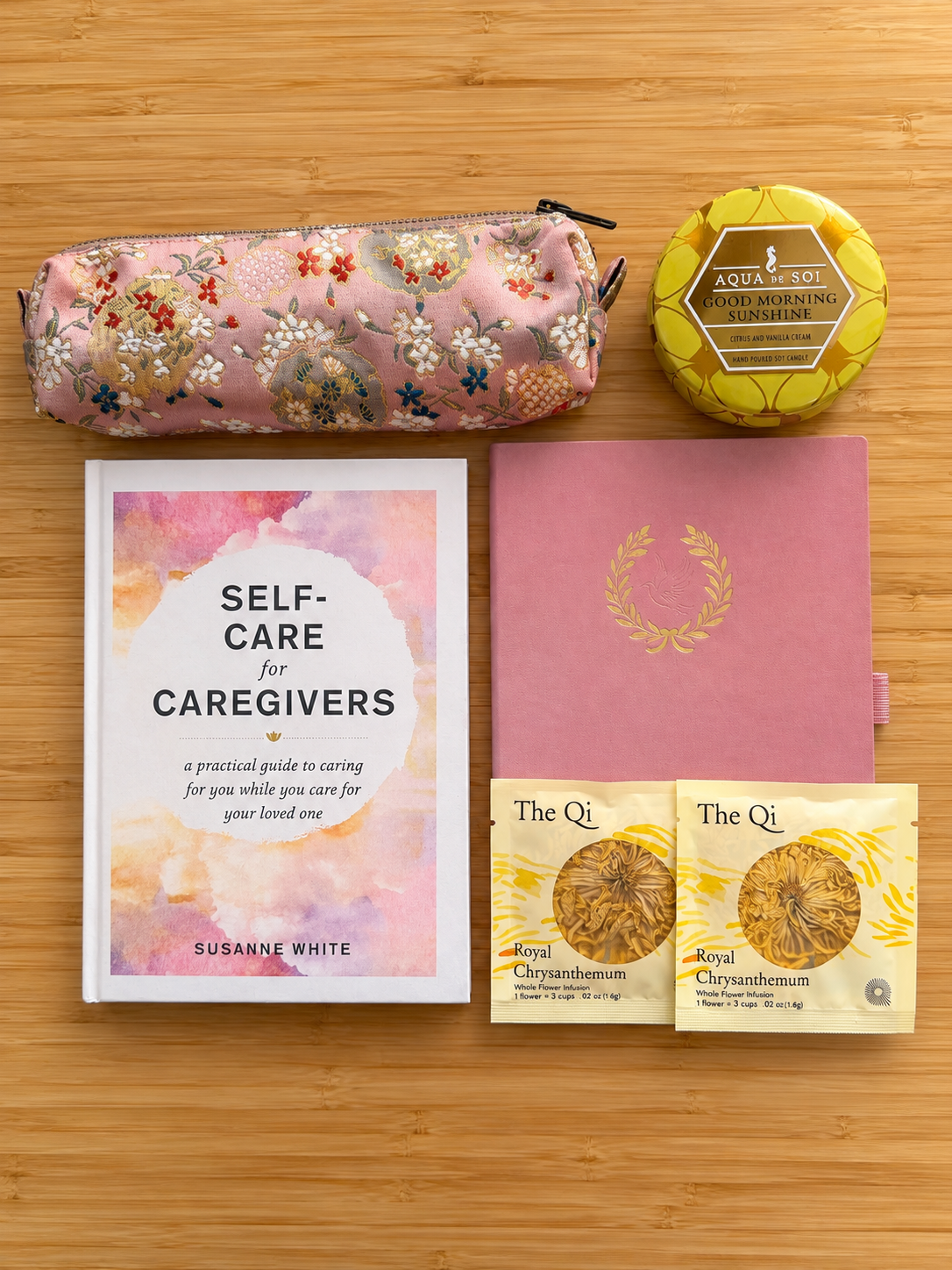 A curated self-care and caregiving gift set arranged on a light bamboo background, including a pink floral zip pouch, a round yellow tin candle, a book titled 'Self-Care for Caregivers' by Susanne White, a pink notebook with gold foil detailing, and two yellow tea bags from The Qi.