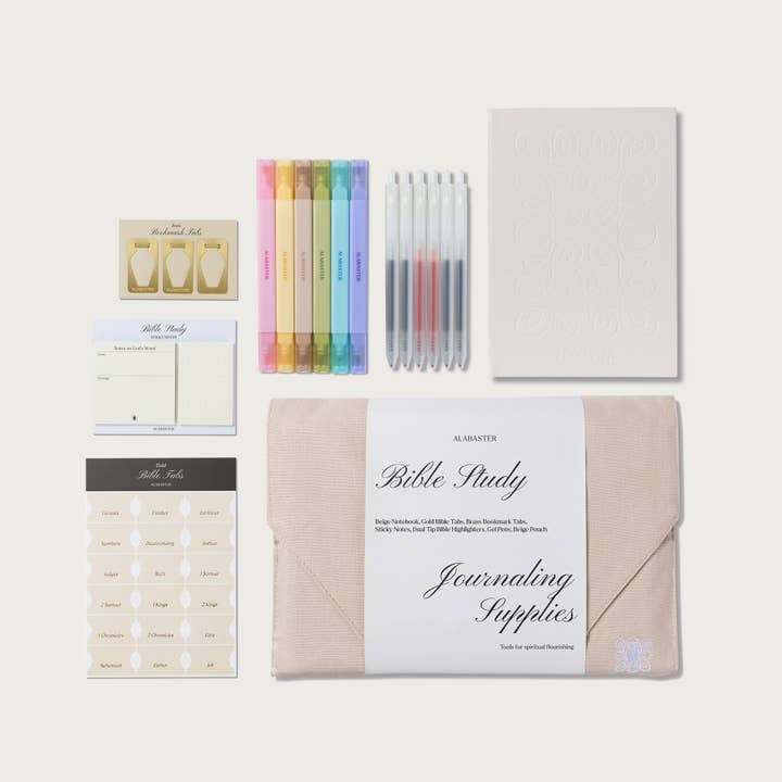 A Bible Study journaling supplies kit by Alastair, arranged neatly on a light beige background. The kit includes a beige envelope, a white paper band with 'Bible Study Journaling Supplies' written on it, a stack of cream-colored sticky notes, a sheet of gold foil tabs, a pastel highlighter pen set, a set of clear pens with colored ink, and a blank embossed journal page.