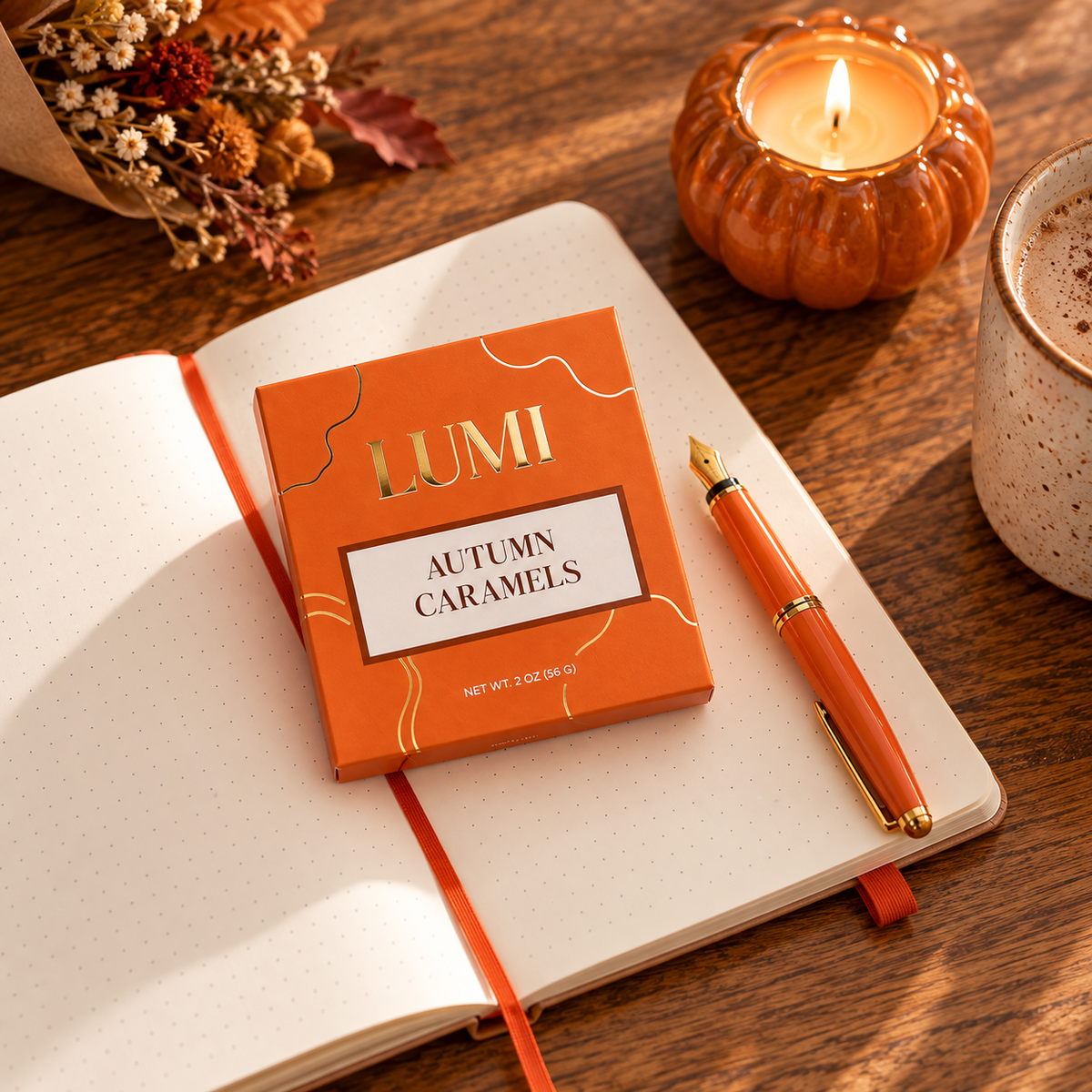 An orange box of LUMI Autumn Caramels rests on an open notebook on a wooden table, styled with an orange pen, a lit pumpkin-shaped candle, and a ceramic mug.