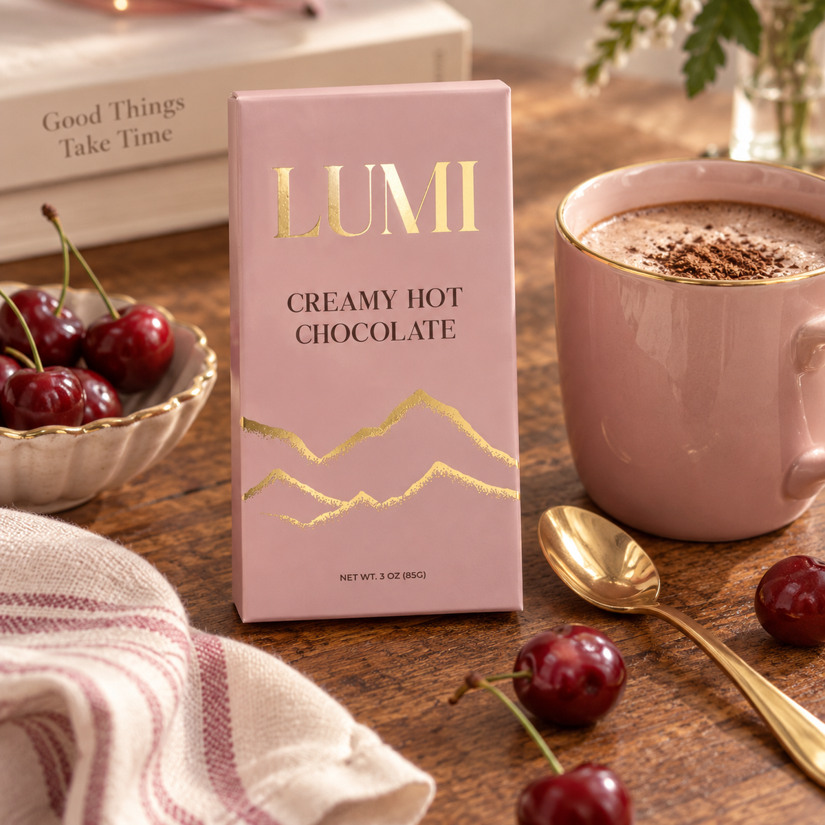 A pink box of LUMI Creamy Hot Chocolate stands on a wooden table, surrounded by a pink mug of hot chocolate, cherries, a spoon, and a striped cloth. The box features gold foil lettering and mountain graphics.