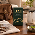 A box of Lumi Mint Hot Chocolate chocolate is displayed on a wooden table in a cozy, styled setting with a brown leather bag, a white marble travel tumbler, a newspaper, and a small dish of chocolate pieces.