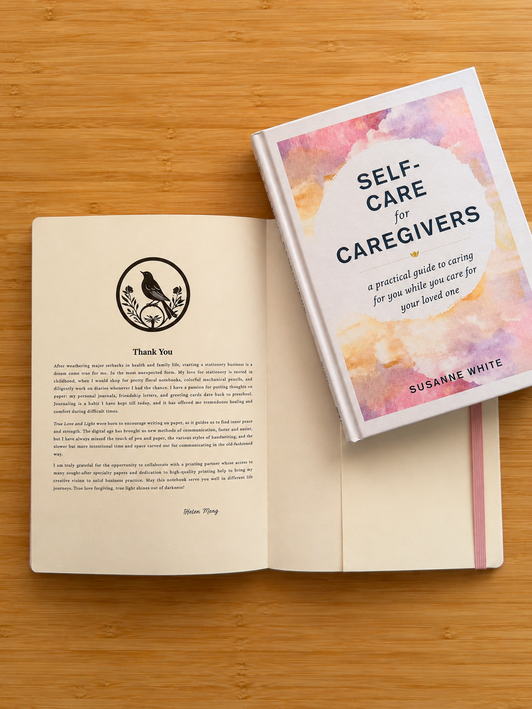 An open book on a bamboo surface, with the front cover titled "SELF-CARE for CAREGIVERS" by Susanne White resting on top. The cover has a pastel watercolor design in pink, yellow, and blue tones, and the open pages show a thank-you message with a bird logo.