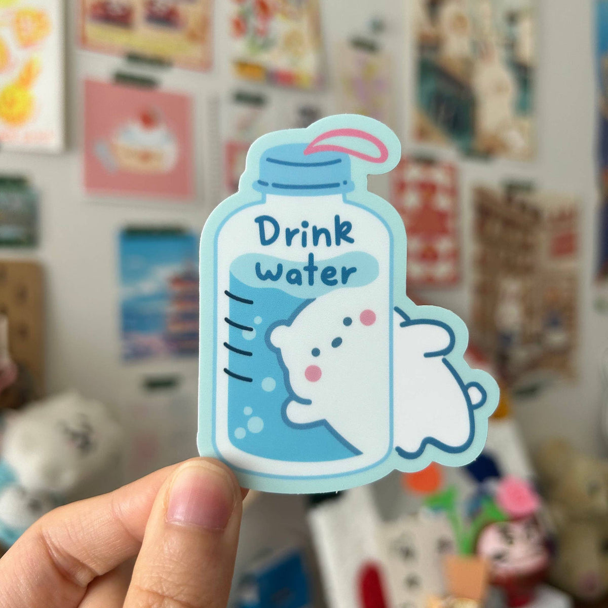 A hand holds a die-cut sticker shaped like a water bottle with the words 'Drink water' and a cute white cartoon character inside, against a blurred indoor background with colorful cards and objects.