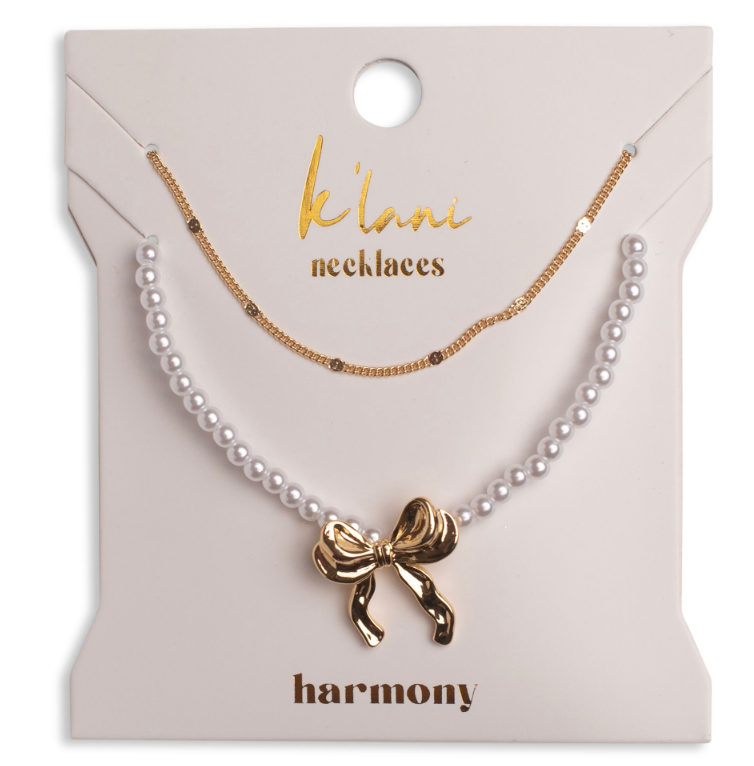 A jewelry gift set on a cream-colored card labeled 'k'lani necklaces' and 'harmony', featuring a white pearl necklace with a gold bow pendant and a delicate gold chain necklace.
