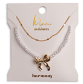 A jewelry gift set on a cream-colored card labeled 'k'lani necklaces' and 'harmony', featuring a white pearl necklace with a gold bow pendant and a delicate gold chain necklace.