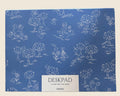 A blue desk pad with a white hand-drawn style pattern of trees, animals, and figures, featuring a white label at the bottom that reads 'DESKPAD', 'PLAN FOR THE WEEK', and 'PAPER'.