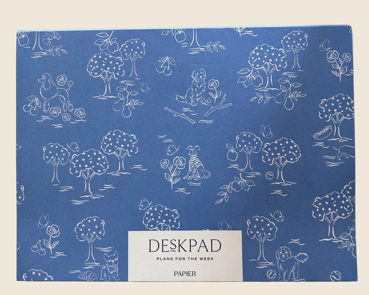 A blue desk pad with a white hand-drawn style pattern of trees, animals, and figures, featuring a white label at the bottom that reads 'DESKPAD', 'PLAN FOR THE WEEK', and 'PAPER'.