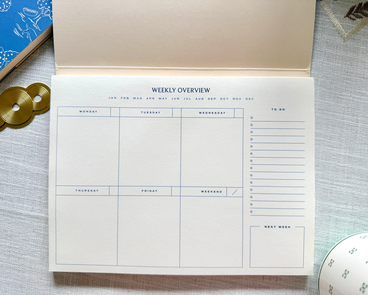 An open weekly planner pad on a textured light gray fabric surface, with gold coins and a blue patterned notebook partially visible as styled props.