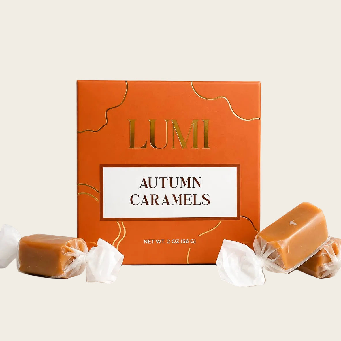 Lumi Autumn Caramels in an orange box with gold lettering, with several unwrapped caramel candies wrapped in white paper scattered around it on a light beige background.