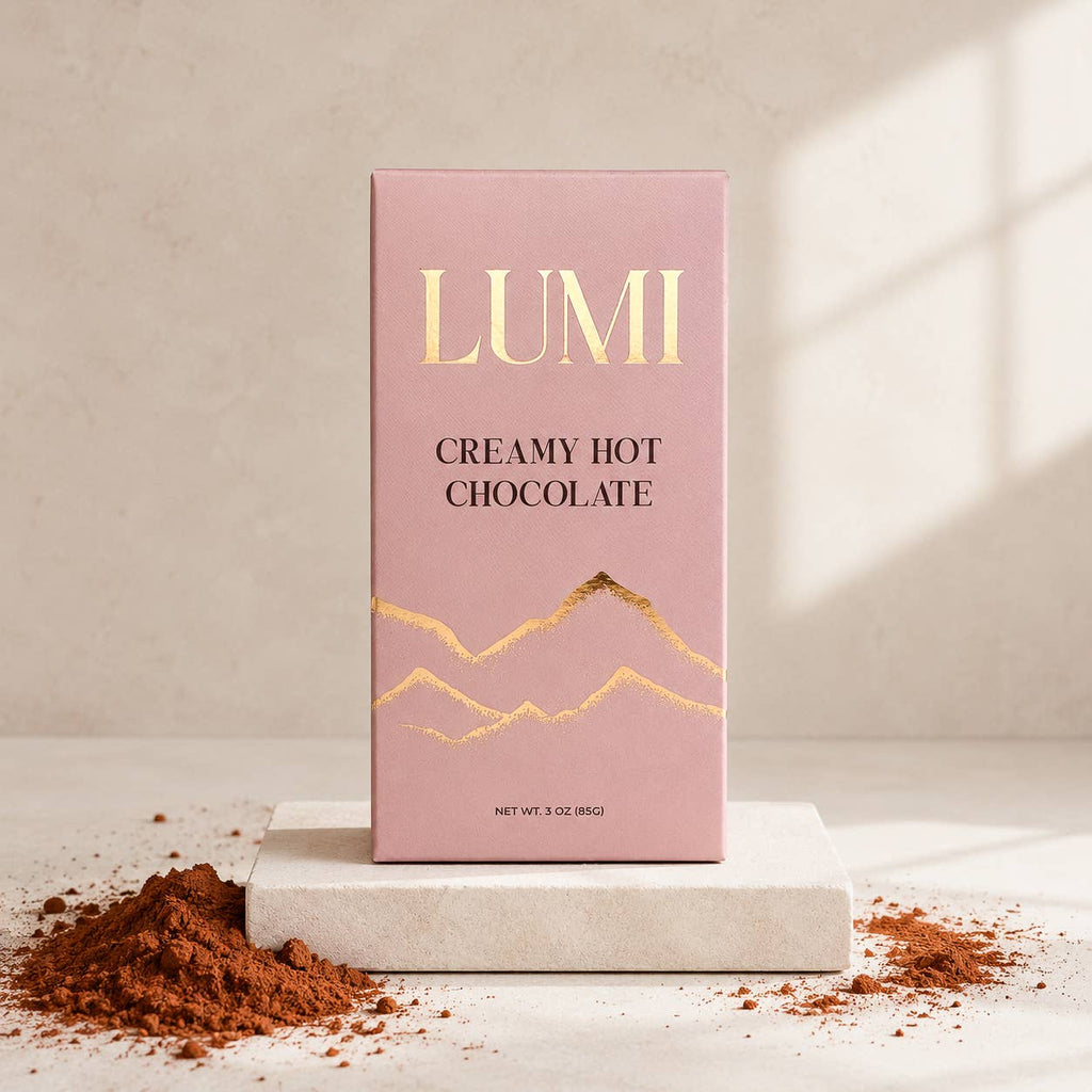 A pink rectangular box of LUMI Creamy Hot Chocolate stands on a white stone slab with cocoa powder scattered around it. The box has gold foil mountain graphics and the brand name LUMI in gold at the top.