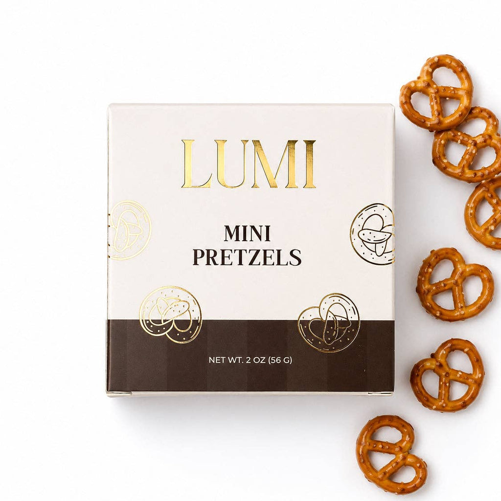 A square white and dark brown box of LUMI Mini Pretzels with gold foil lettering, shown on a white background with several pretzels scattered to the right.