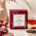 A box of LUMI Wild Cranberry Black Tea is displayed on a light stone surface with a glass of brewed tea, loose tea leaves, and cranberries in a warm, rustic setting.