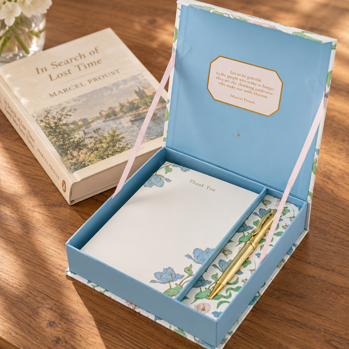 A floral gift box set on a wooden table, featuring a light blue box with a pink ribbon, a floral notepad, a gold pen, and a book titled 'In Search of Lost Time' by Marcel Proust in the background.