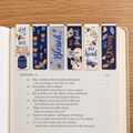 Six decorative book bookmarks with floral and bee illustrations are arranged on an open Bible page, with the text Proverbs 31 visible underneath on a wooden tabletop.