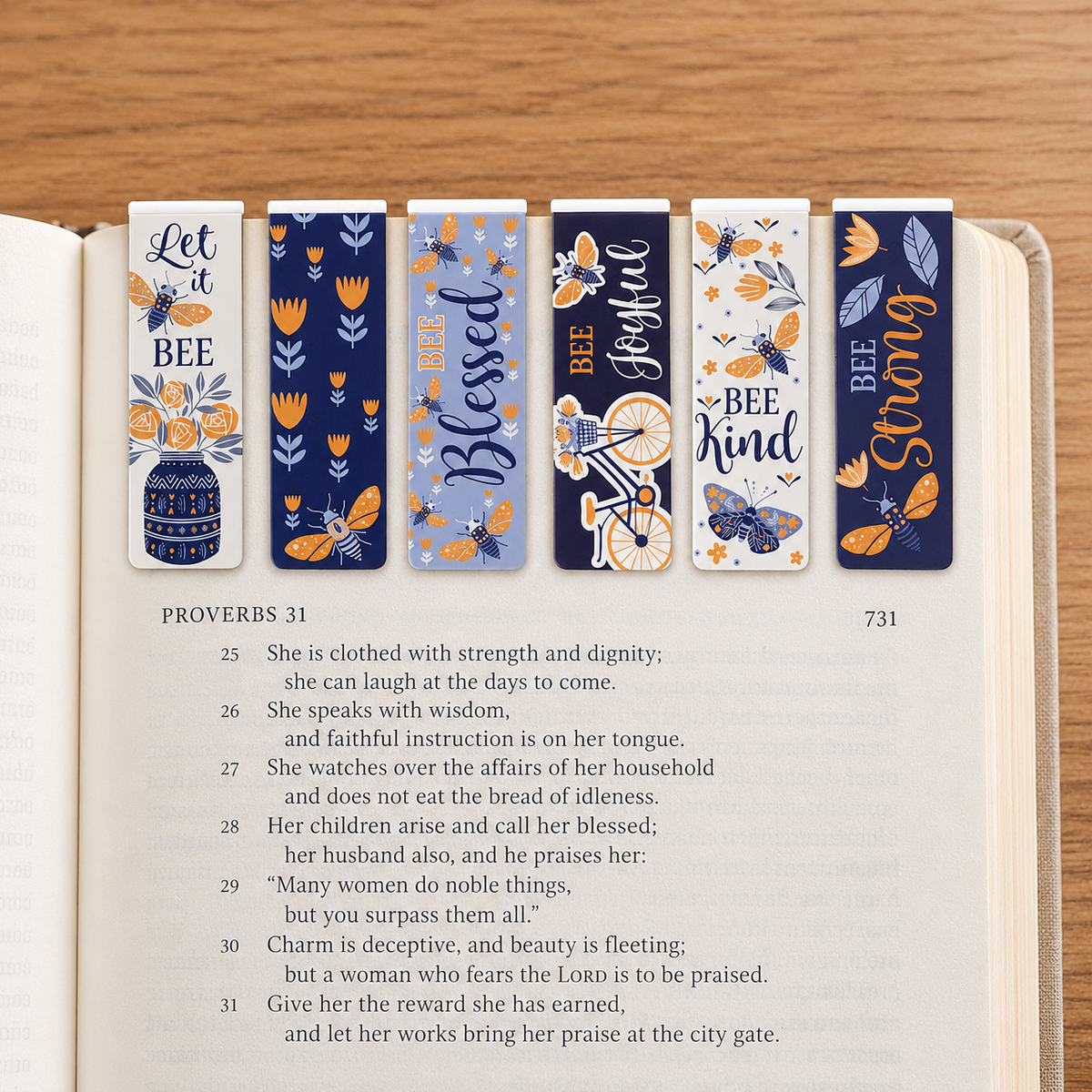 Six decorative book bookmarks with floral and bee illustrations are arranged on an open Bible page, with the text Proverbs 31 visible underneath on a wooden tabletop.