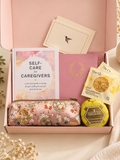 A curated gift box set for caregivers, displayed in an open light pink box. The set includes a 'Self-Care for Caregivers' guide, a pink floral zippered pouch, two yellow tea bag packet, and a tin candle, with a card featuring a hummingbird in the background.