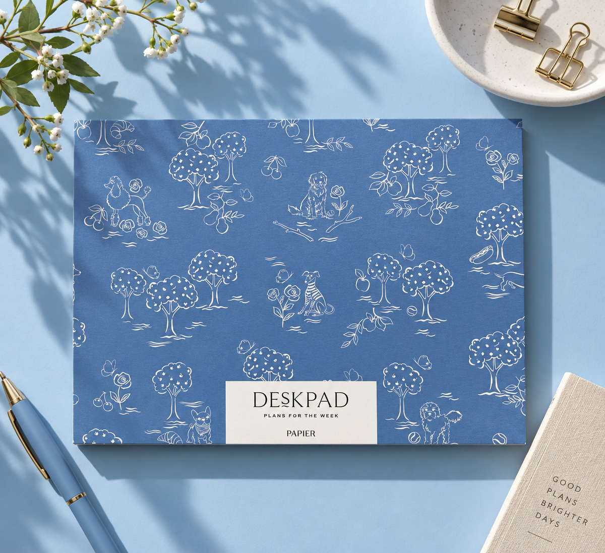 A blue desk pad with a white line-art pattern of trees, animals, and small doodles, styled on a light blue surface with a pen, paper clips, and a notebook nearby.