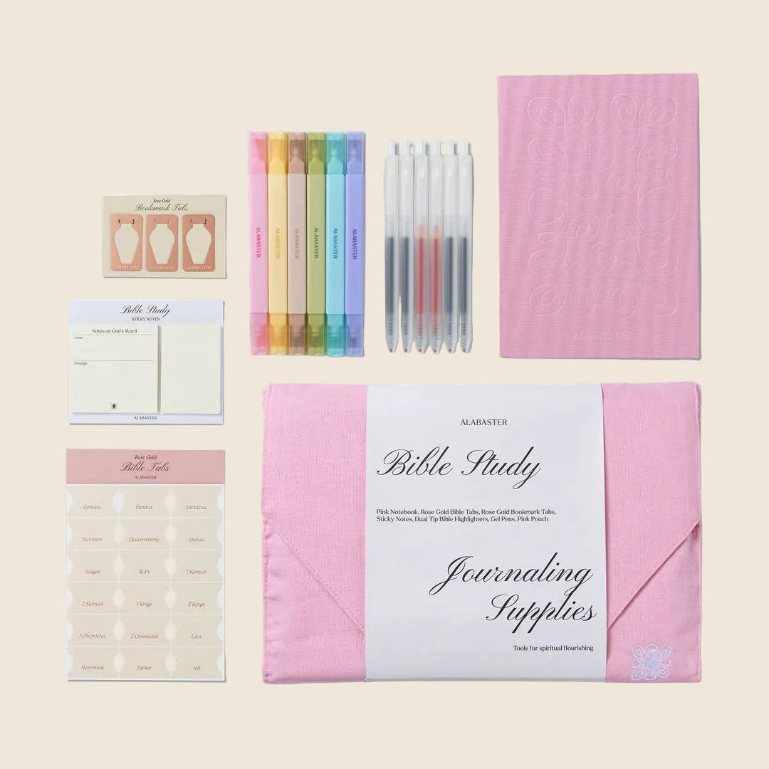 A Bible study journaling supplies kit by Alabaster Co., arranged on a light beige background, including a pink journal, matching pink envelopes, colored highlighter pens, gel pens, sticker sheets, and notepad pages.