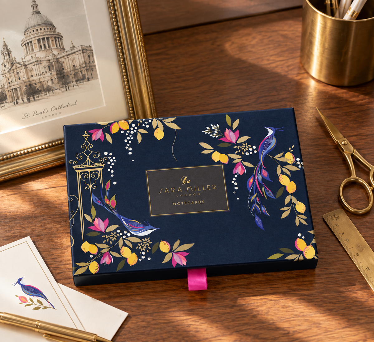 A navy blue Sara Miller London notecard box with floral and bird illustrations, styled on a wooden desk with gold scissors, a brass cup, and a framed print.