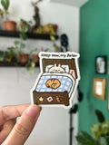 A hand holds a die-cut sticker featuring a cute cartoon bunny sleeping in a bed with the text 'sleep now, cry later'. The background shows a softly blurred indoor room with shelves of plants and a green wall.