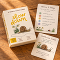 The 'slow down' mindful activity cards set by Magic Cat, featuring a cream-colored box and two sample cards displayed on a warm wooden tabletop. The cards feature yellow title text, black instructions, and illustrations of snails, ladybugs, and flowers.