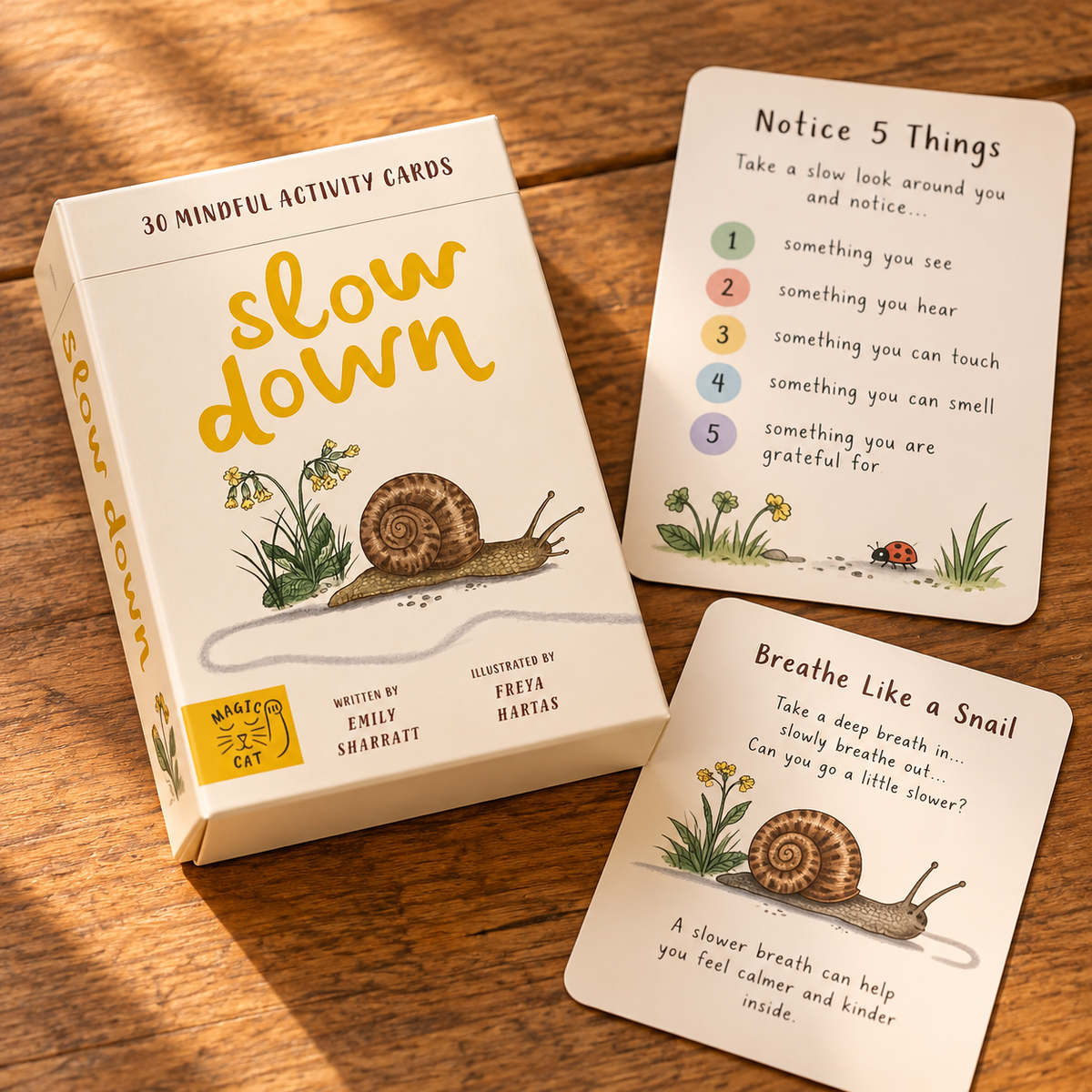 The 'slow down' mindful activity cards set by Magic Cat, featuring a cream-colored box and two sample cards displayed on a warm wooden tabletop. The cards feature yellow title text, black instructions, and illustrations of snails, ladybugs, and flowers.