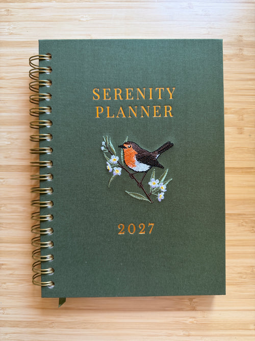 Showing two spiral-bound 2027 Serenity Planner, one with a dark green cover and the other one a light peach cover, both featuring a robin bird and white flowers illustration, placed on a light wood background.