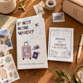 Two travel-themed notebooks displayed on a wooden desk surrounded by travel accessories, including a pen, stamps, tape, a pencil case, and a mug.