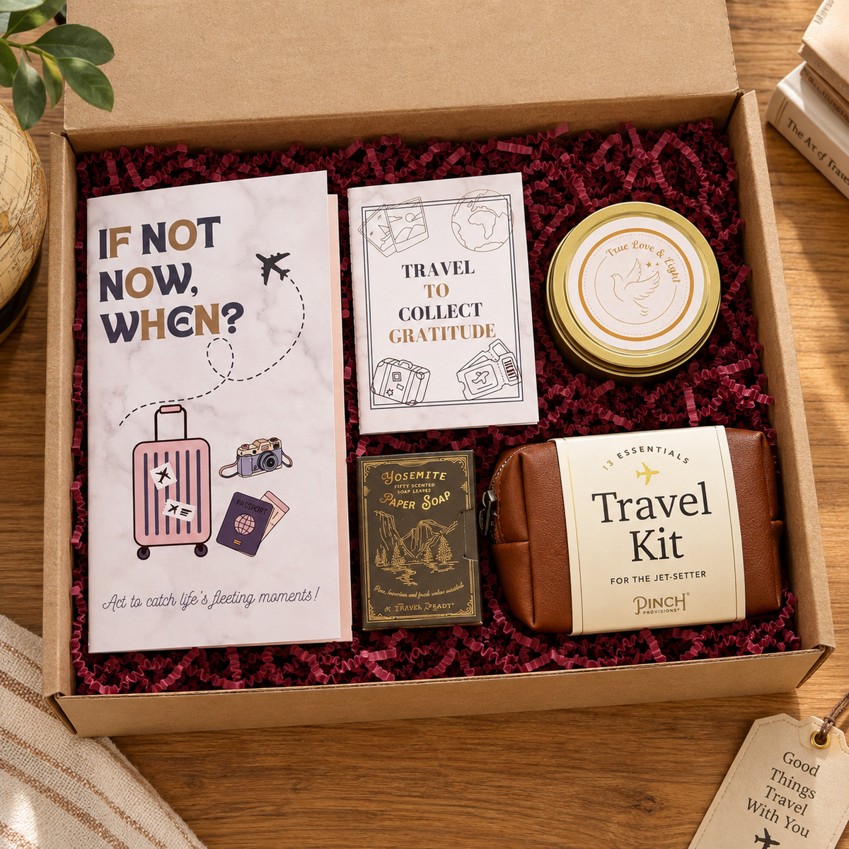 A curated travel-themed gift box set from Amazing Robin Gifts, arranged inside an open brown cardboard box filled with burgundy shredded paper. The set includes a white card reading 'IF NOT NOW, WHEN?', a 'Travel to Collect Gratitude' card, a round tin labeled 'The Great Gratitude Bake', a small brown leather pouch labeled 'Travel Kit', and a olive green pack of paper soap.