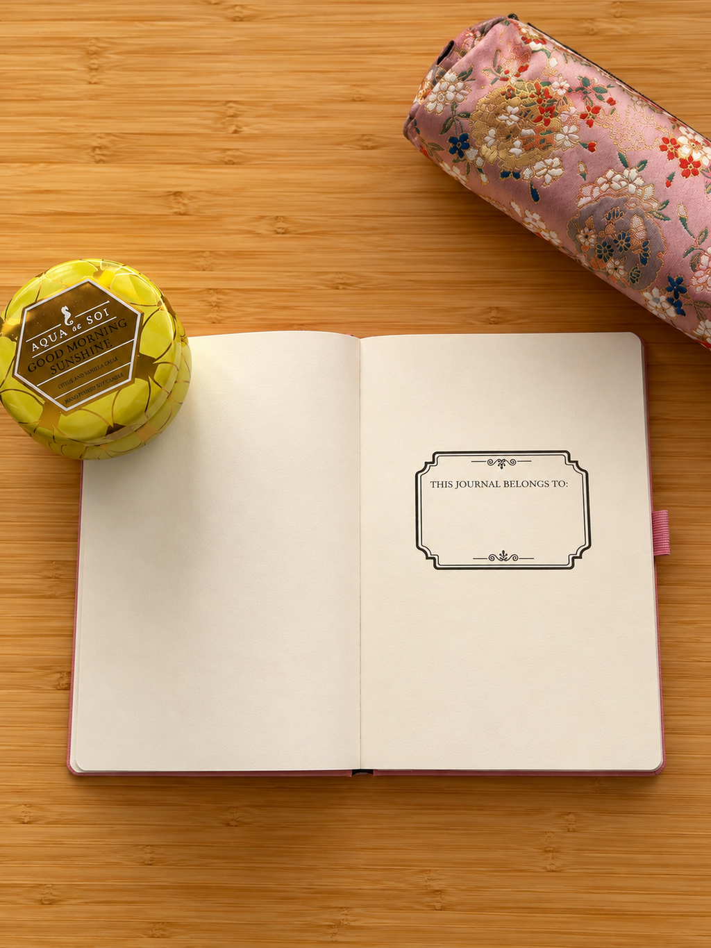 An open pink journal with blank cream pages lies on a light wood surface, alongside a yellow tin candle and a pink floral pencil case.