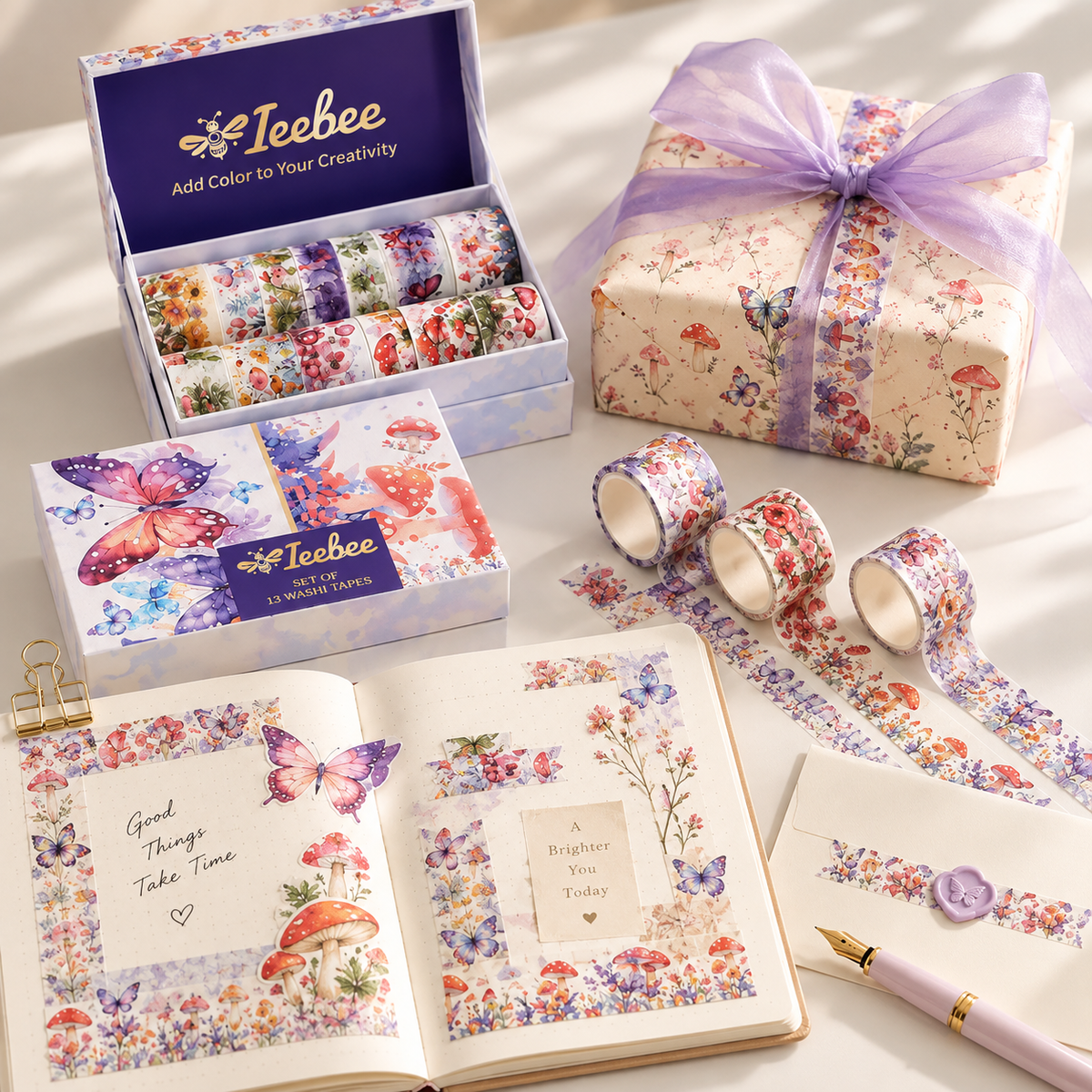 A coordinated stationery gift set arranged on a light fabric surface, including an open notebook with floral washi tape borders, a sealed envelope with a purple seal and floral decoration, a wrapped gift box with a lavender ribbon, an open box of patterned washi tape rolls, and two loose rolls. The products show the brand name Ieebee on the packaging.